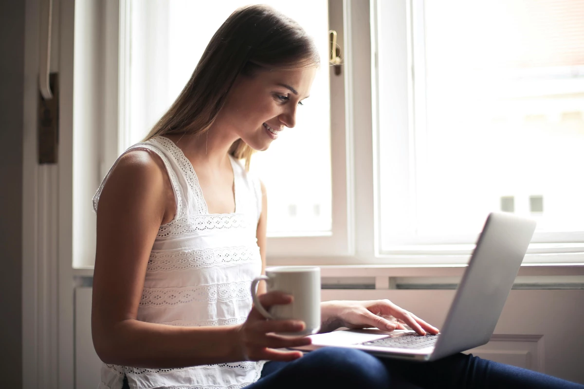 A woman smiling while using a laptop at home, with a cup of coffee nearby